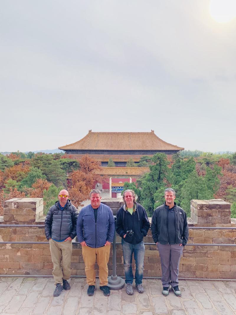 Four men pose on a brick terrace at the Ming Tombs, a golden-roofed hall rising behind autumn trees