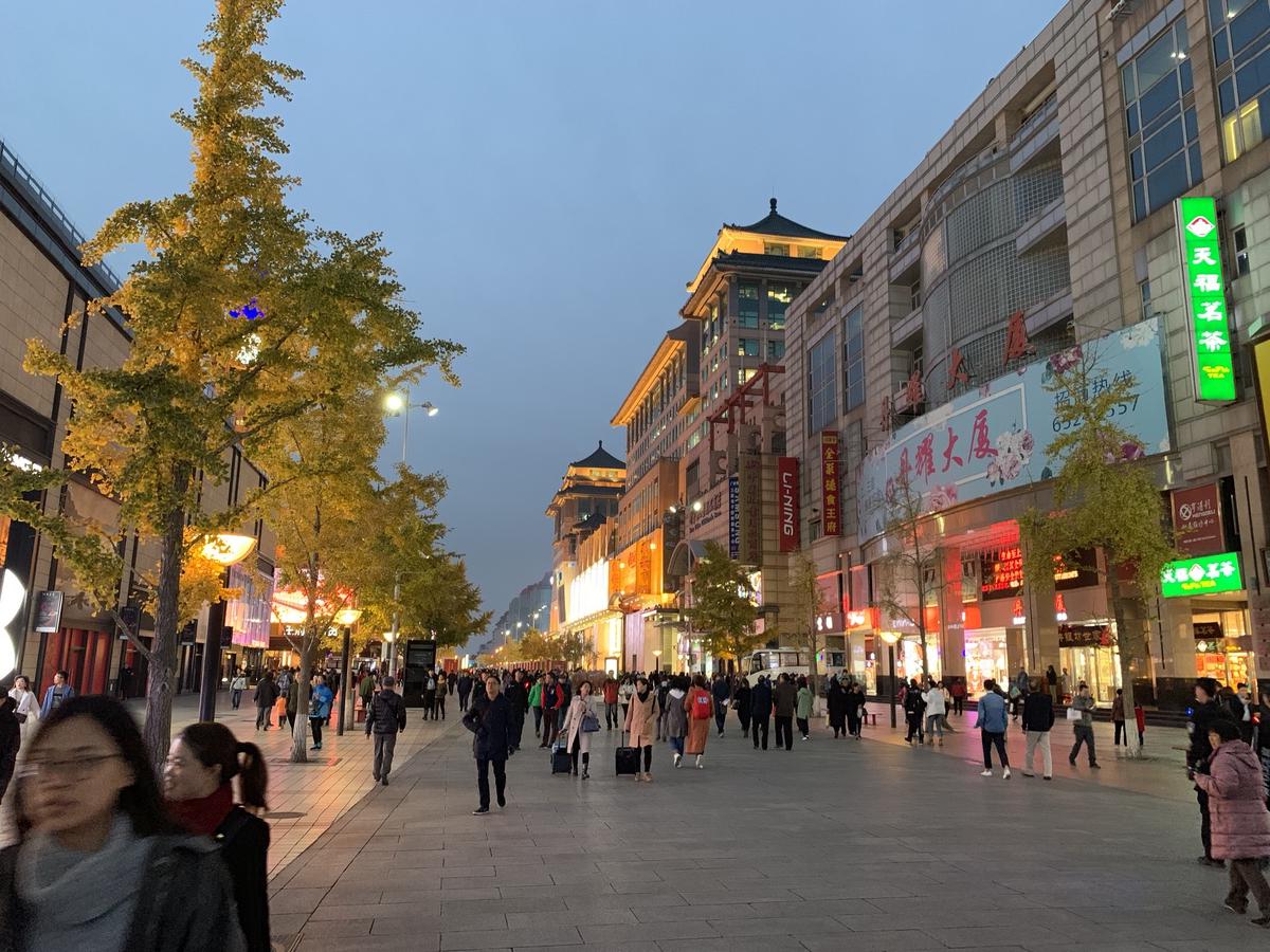 Shoppers stroll the Wangfujing pedestrian street at dusk beneath lit signs and yellow ginkgo trees