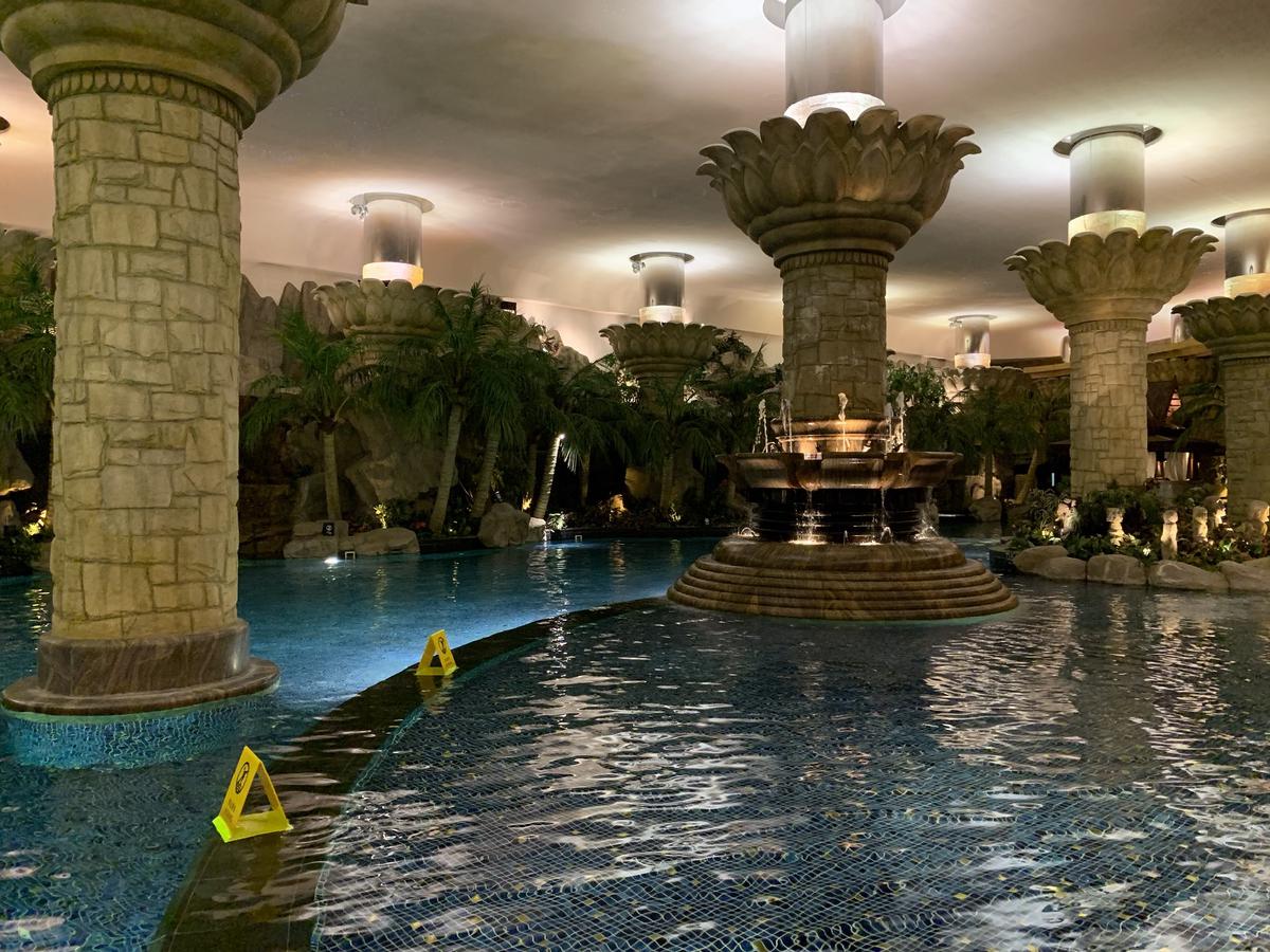 Indoor hotel pool with lotus-topped stone columns, a tiered fountain, and palm trees under warm lights