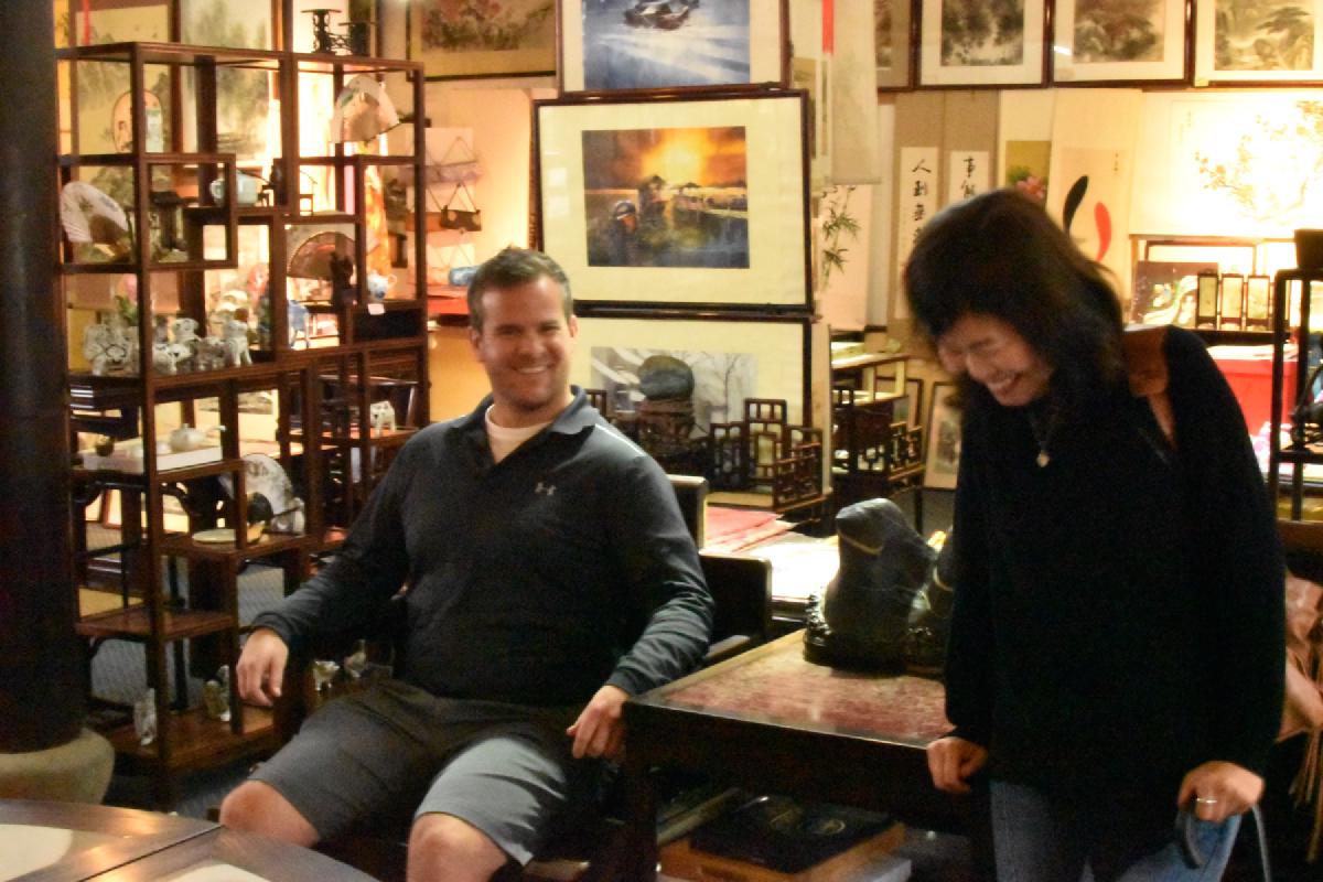 A man sits grinning in a carved wooden chair while a woman laughs beside him, surrounded by scroll paintings in a Shanghai art shop