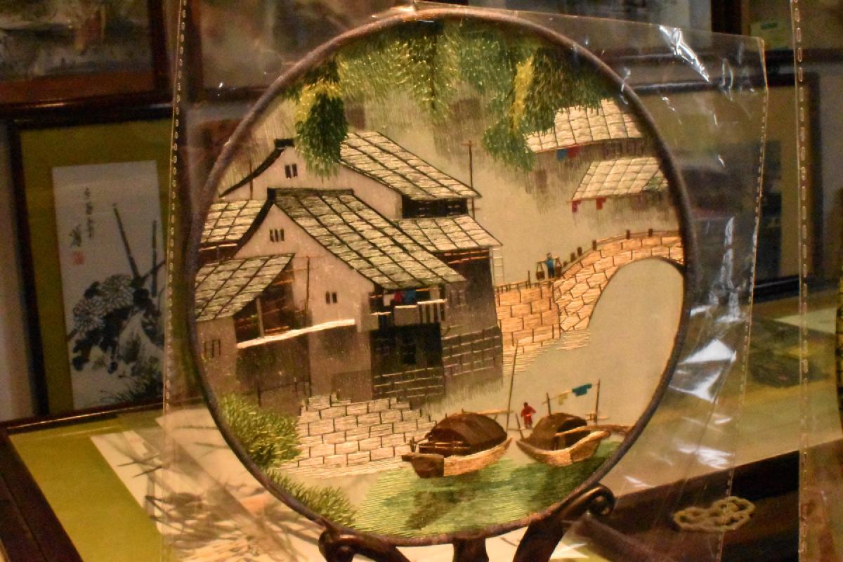 A round silk embroidery of a water town, with tiled houses, an arched stone bridge and two boats, displayed on a stand for sale