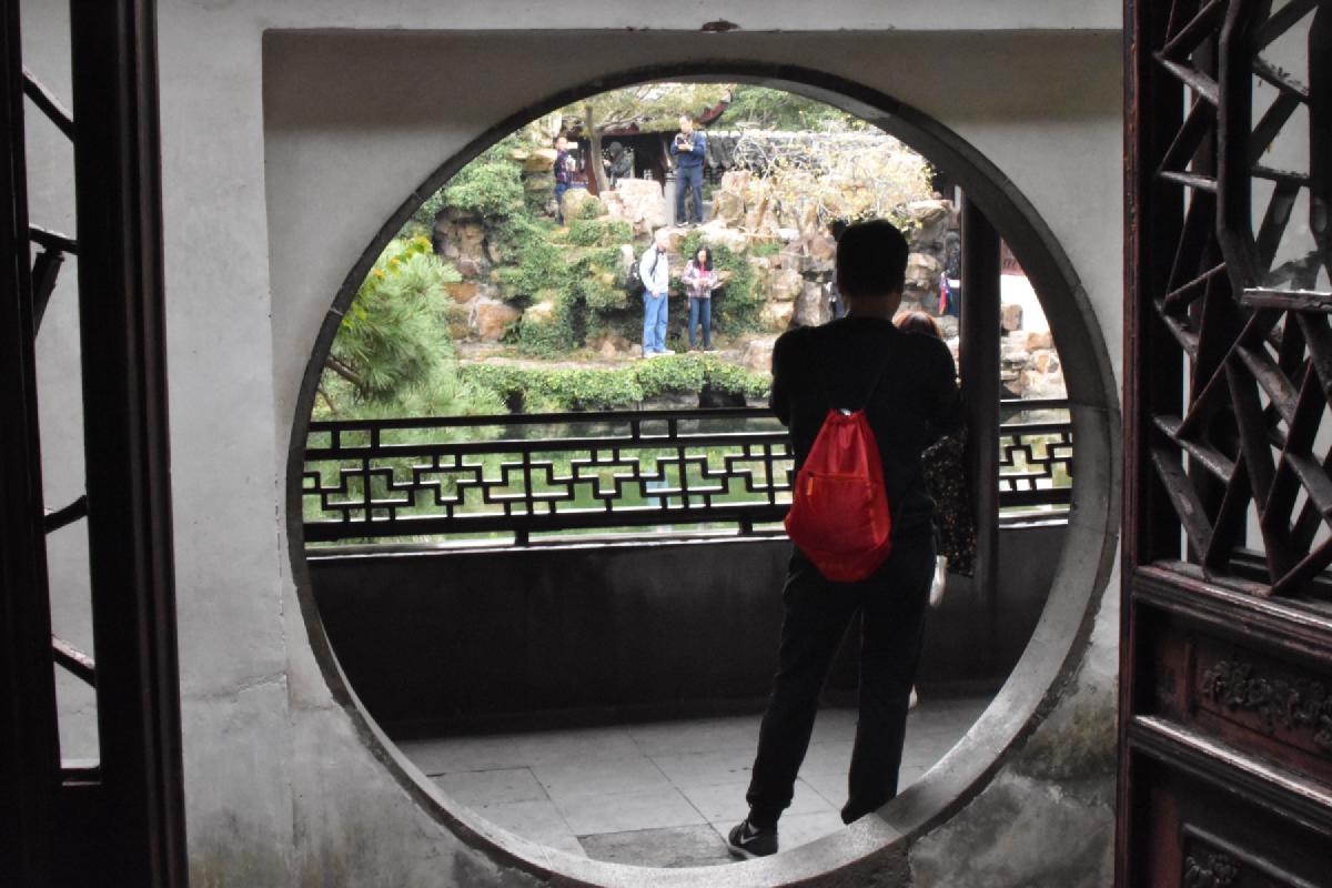 A visitor with a red drawstring bag stands in a circular moon gate, looking out at tourists climbing a garden rockery in Shanghai