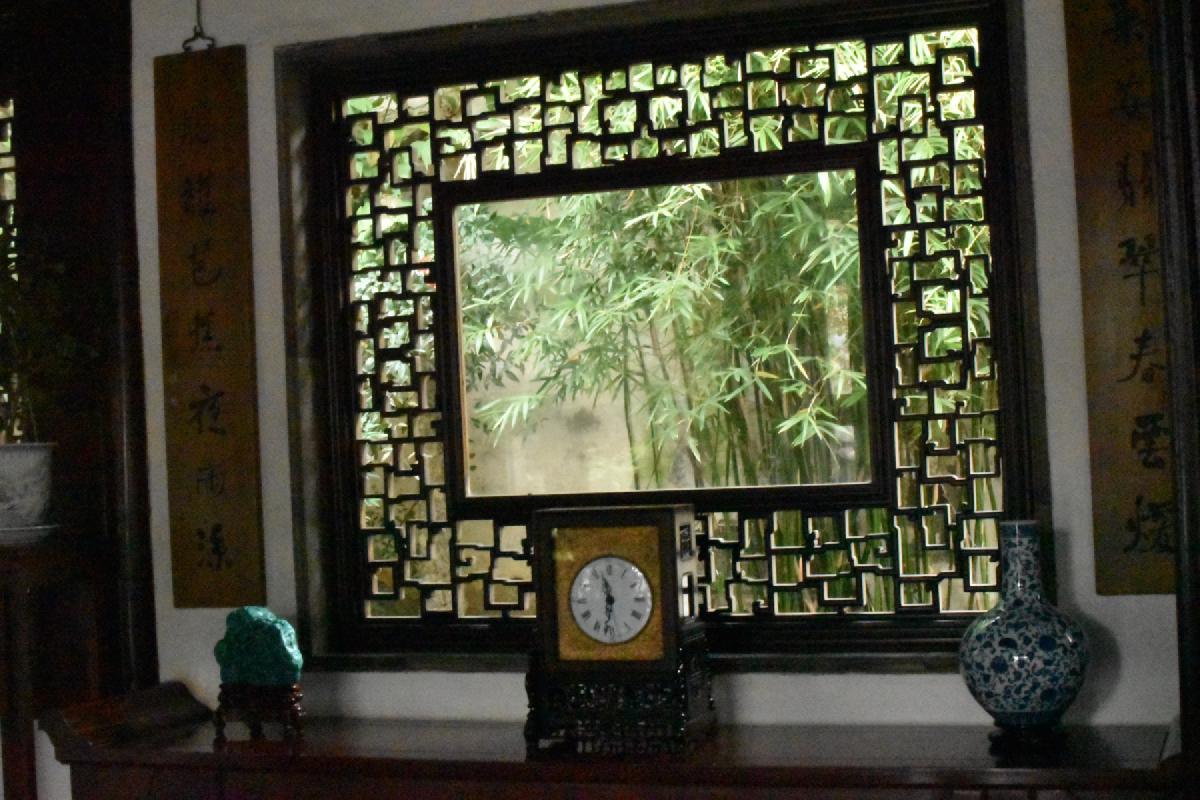 A mantel clock, blue-and-white vase and green stone sit below a latticed window looking onto bamboo, flanked by calligraphy scrolls