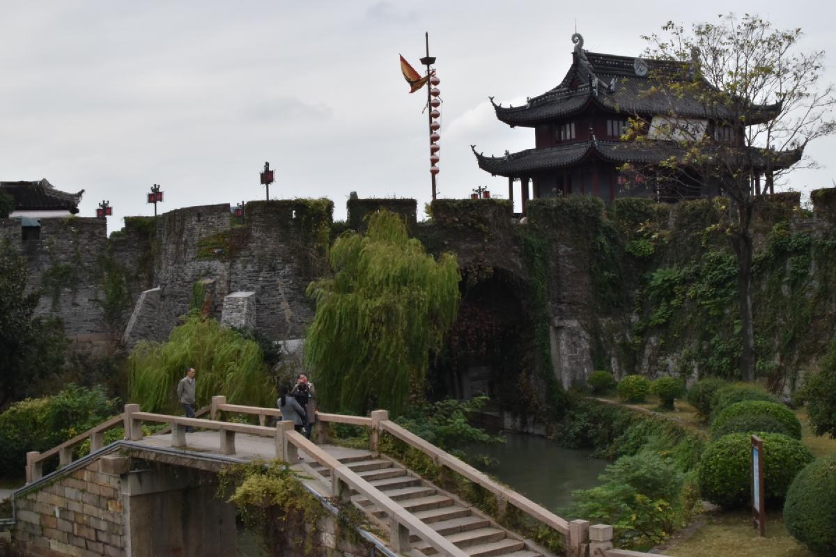 Visitors on a stone arch bridge below an ivy-covered city wall and tiered wooden gate tower in China