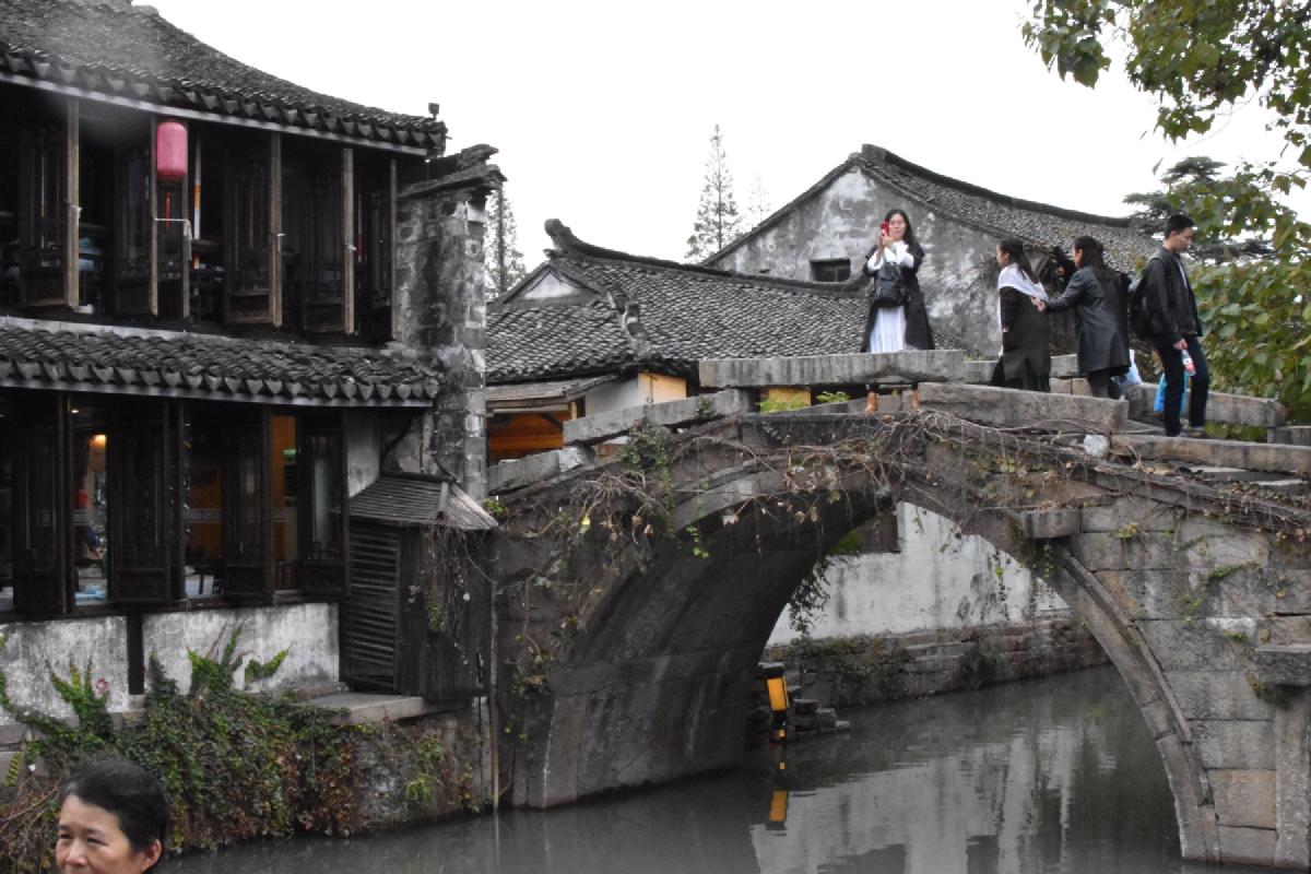 People crossing a stone arch bridge over a canal beside tiled-roof wooden houses in a Chinese water town