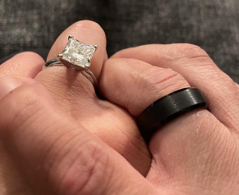 Two clasped hands, a princess-cut solitaire on one ring finger and a wide brushed black band on the other